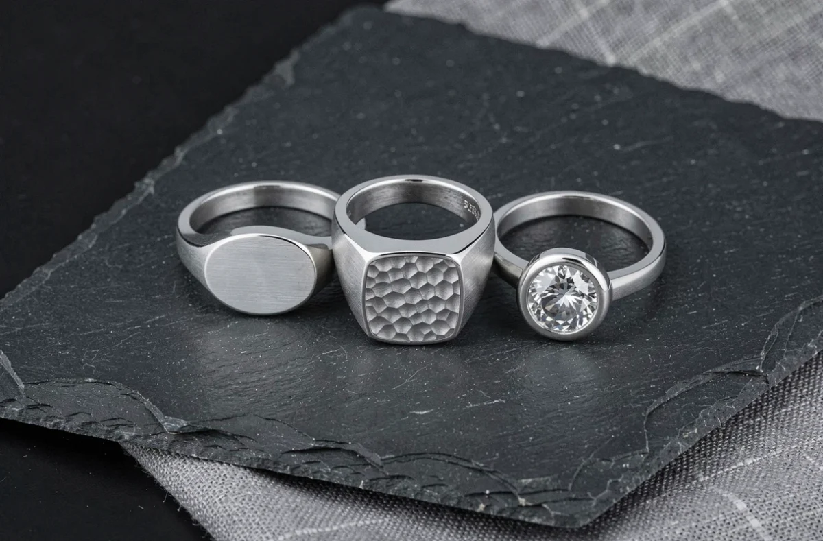 Trois chevalières en or blanc pour homme présentées sur ardoise noire avec différents designs : ovale, carré martelé et rond avec diamant